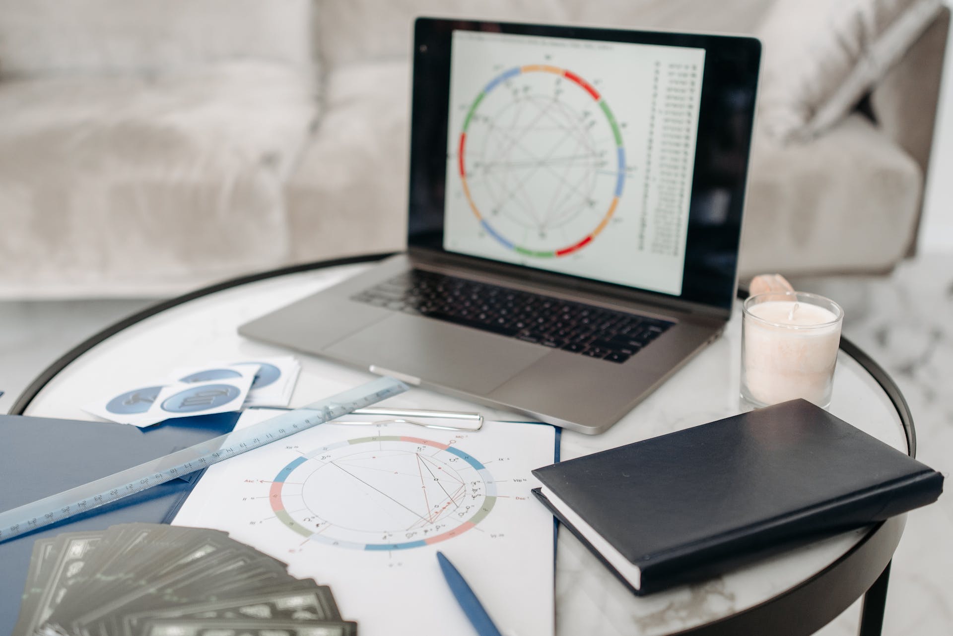 Photo of laptop with a birth chart on the screen. Chart printouts on desk with Zodiac cards, ruler, notebook, and glass of milk.